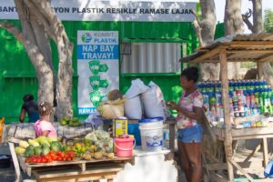 zu sehen ist ein Marktstand in Haiti. Eine Frau hat mehrere Beutel Plastikmüll vor sich stehen. Links im Bild ist viel Obst auf einem Tisch aufbereitet. Rechts sind verschiedene Getränke und Kosmetikprodukte für den Alltag. Im Hintergund hängt ein großes Schild, welches zeigt, dass man Plastikflaschen gegen Geld oder verschiedene Gegenstäde eintauschen kann